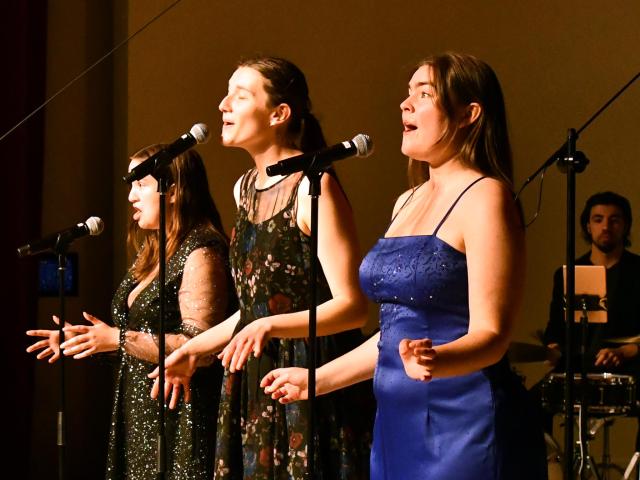 Three women sing into microphones on stage; the first wears a black sparkly dress, the second a floral dress, and the third a blue gown. A drummer in the background plays a drum set. The setting has a warm, dimly lit atmosphere.