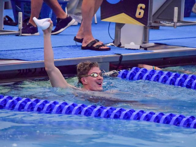 A swimmer in goggles and swim cap raises a hand triumphantly while standing in a pool lane. Another swimmer is nearby. Blue lane dividers and a diving block marked "6" are visible. Spectators' legs and sandals can be seen on the pool deck.