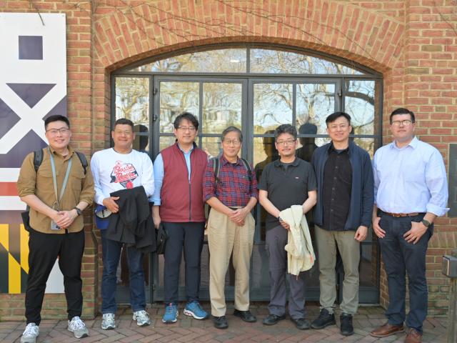 Seven people stand side by side in front of a brick building with a glass door, smiling at the camera. Some hold jackets. There is a large sign on the left side of the image.