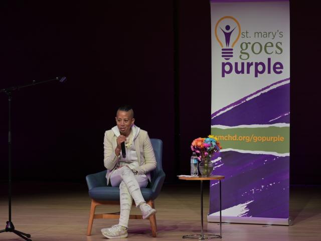 A person sits on a chair speaking into a microphone on stage, next to a banner with "St. Mary's Goes Purple" and a small table with flowers and a water bottle.