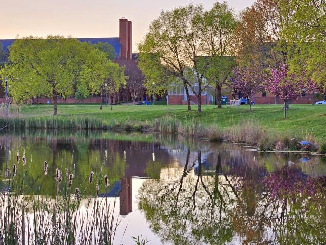 A calm pond reflects trees and a brick building on a grassy campus at sunset, with some cars parked in the background. A great blue heron wades at water's edge.