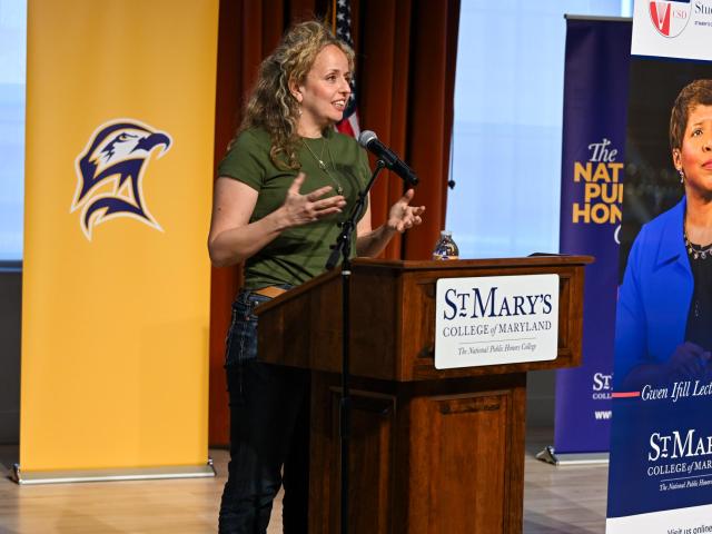 A woman speaks into a microphone at a podium with banners and a sign reading "St. Mary's College of Maryland" in the background.