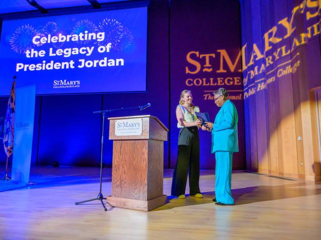 Two people shake hands on stage at St. Mary’s College of Maryland event, with a screen reading "Celebrating the Legacy of President Jordan" in the background.