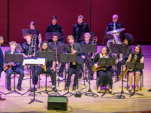 A jazz band with 12 musicians performs on stage with various instruments like saxophones, trombones, and a tuba. A conductor stands in front, and music stands are visible.