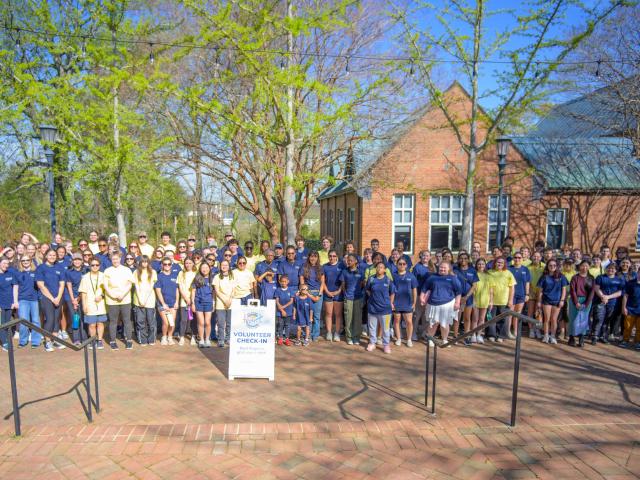 A large group of people wearing yellow and blue shirts stand outside on a brick path near trees, with a sign reading "Volunteer Check-In" in front.