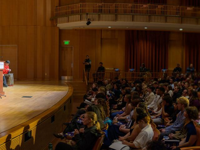 A speaker addresses an audience from a podium on a stage, with officials seated nearby in academic regalia. The auditorium is filled with attendees.