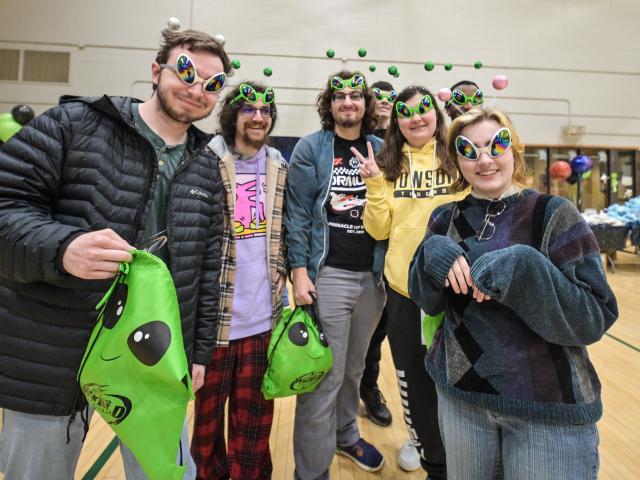 Five people wearing alien-themed headbands and glasses, holding green bags with alien faces, stand indoors on a gym floor, smiling at the camera.
