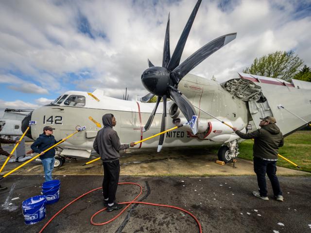 Several people wash a large military aircraft with mops and brushes outdoors, with cleaning supplies and buckets visible around them.