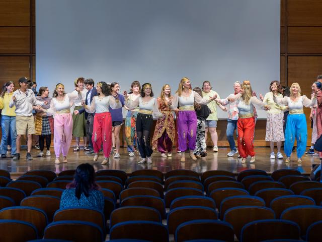 A group of people perform a group dance on a stage in front of an audience in a wooden auditorium.