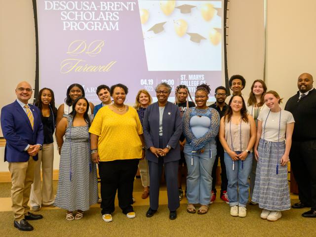 A group of students and staff pose for a photo in front of a screen displaying "Desousa-Brent Scholars Program" during an indoor event.