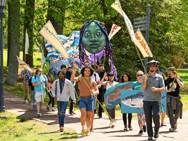 A group of people march outdoors carrying banners, a large green face puppet, and a fish sign on a sunny day in a park-like setting.