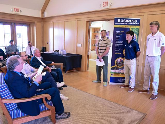Three young men stand and present to a seated group in a wood-paneled room, with a business program banner visible in the background.