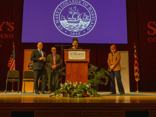 Four people stand on stage at a St. Mary’s College of Maryland event, with a speaker at a podium and the college seal projected in the background.