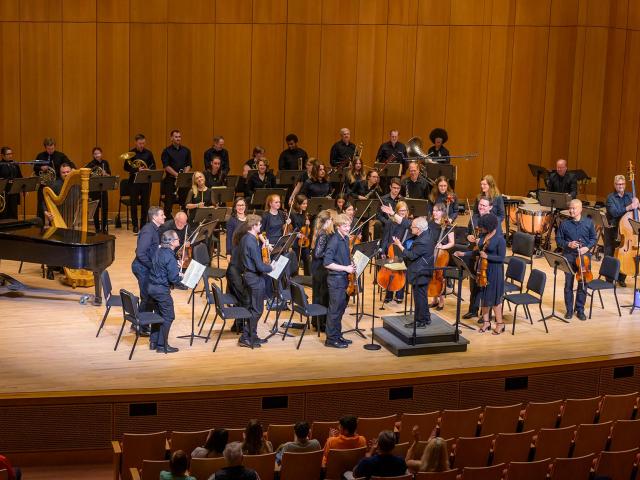 An orchestra performs on stage in a concert hall, with musicians playing various instruments including strings, woodwinds, brass, harp, and piano. Audience members are seated in front.