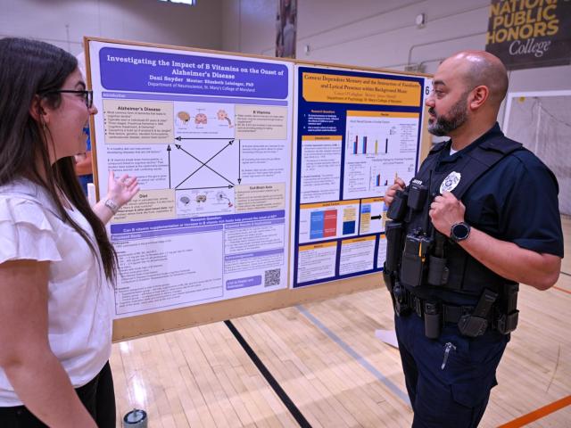 A woman explains a scientific research poster to a uniformed police officer in a gymnasium with a volleyball net in the background.