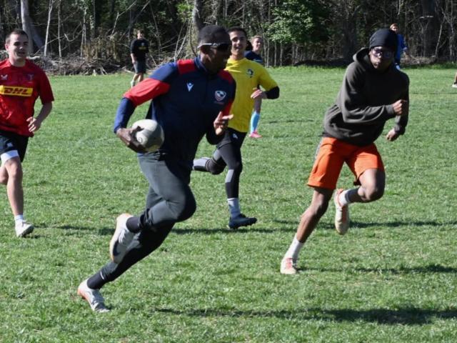 A group of men playing rugby on a grassy field. One player in a dark shirt is running with the ball, pursued by others. Trees and additional players are visible in the background.