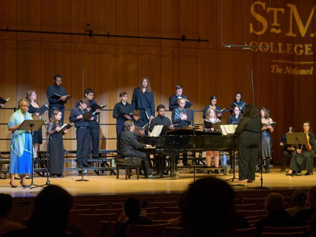 A choir and pianist perform on stage in a concert hall, with a conductor leading and an audience seated in the foreground.
