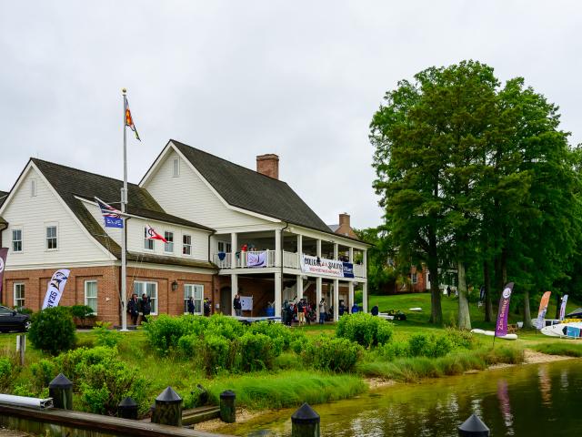 A two-story brick and white building with flags and banners hosts an event near a sandy waterfront, surrounded by greenery and trees.