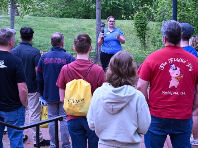 A tour guide speaks to a group of people outside on a brick walkway, with grass and trees visible in the background.