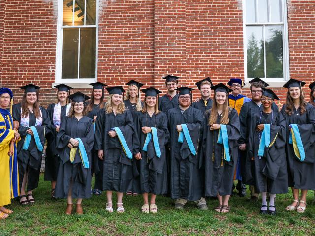 A group of graduates and faculty members in academic regalia pose for a photo in front of a brick building.