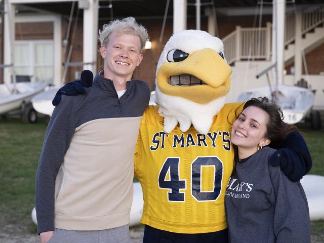 Two students stand smiling next to a person in an eagle mascot costume wearing a St. Mary's jersey numbered 40, with boats and a building in the background.