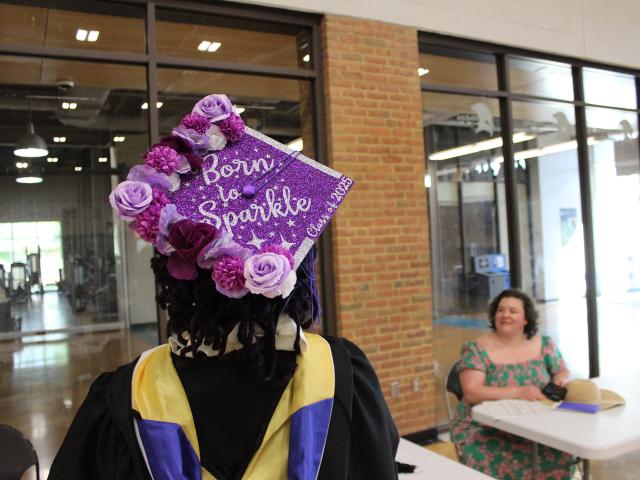 A graduate wearing a decorated cap reading "Born to Sparkle" stands near a table where a woman is seated, inside a building with glass walls and brick accents.