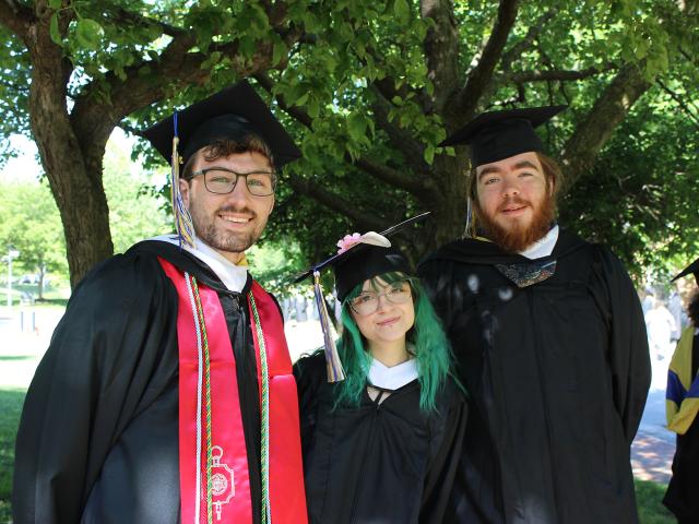 Three graduates in caps and gowns stand outdoors under a tree, smiling at the camera on a sunny day.