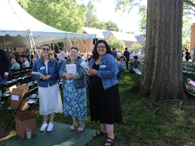 Three women in denim jackets stand and smile near a tree at an outdoor event with chairs and a tent in the background.
