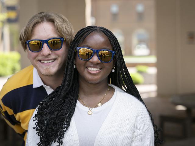 Two people wearing sunglasses are smiling outdoors. The person in front has long braids and white clothing; the person behind has light hair and a striped shirt.