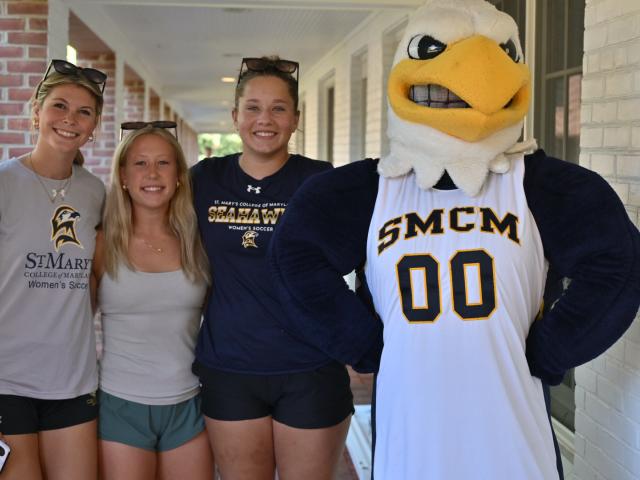 Three young women stand next to a person in a Seahawk mascot costume wearing an "SMCM 00" jersey, posing for a photo outdoors by a brick building.