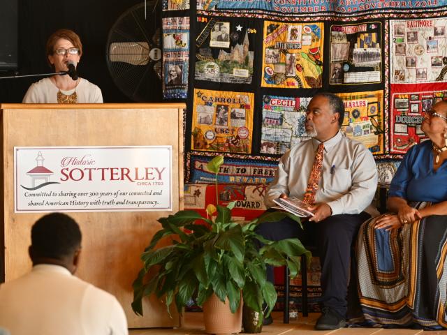 A woman speaks at a podium labeled “Historic Sotterley” while a man and woman sit onstage in front of a colorful quilt backdrop.
