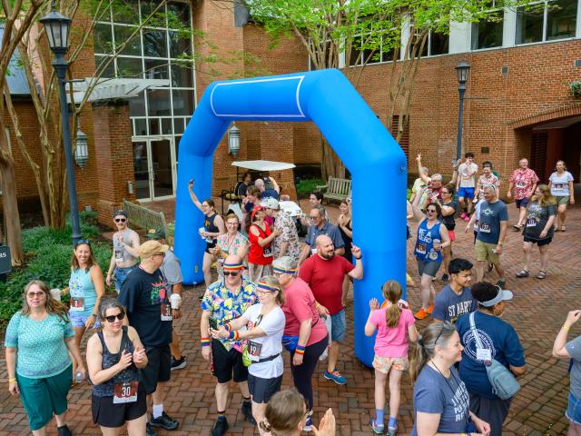 A group of people gathers under and around a blue inflatable arch outdoors, some wearing race bibs, casual clothes, and holding drinks, with a mix of cheering and smiling participants.
