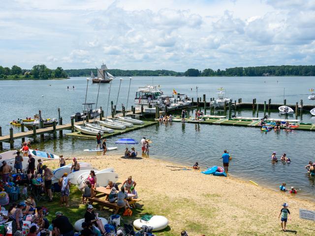Crowd of people at a lakeside beach and marina, with boats docked, people swimming, and groups gathered on the sand and grass under a partly cloudy sky. A 17th-century replica sailing ship sails in the background.