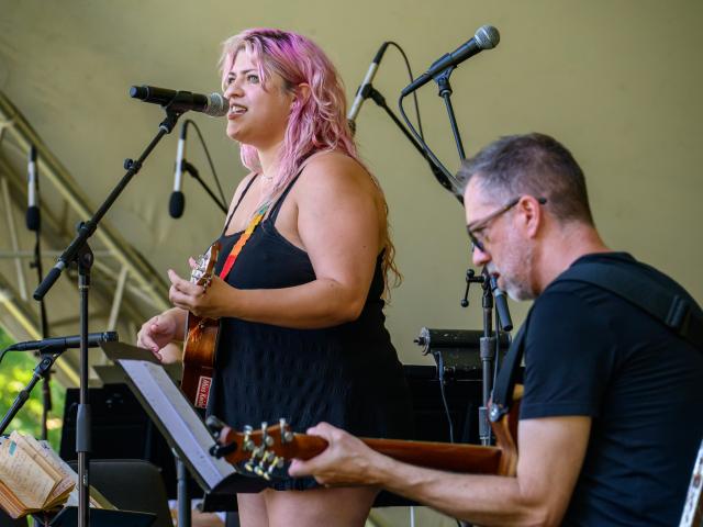 A woman with pink hair sings and plays ukulele on stage next to a man playing guitar. Microphones and music stands are visible in the background.