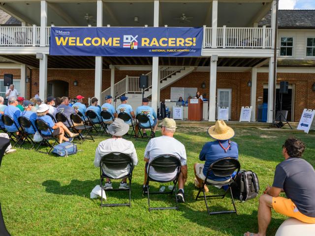 A group of people sit on chairs outdoors facing a stage where a speaker addresses them under a large banner that reads "WELCOME RACERS!"