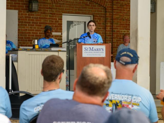 A person speaks at a podium labeled "St. Mary's College of Maryland" during an outdoor event, with people seated and listening in the foreground.