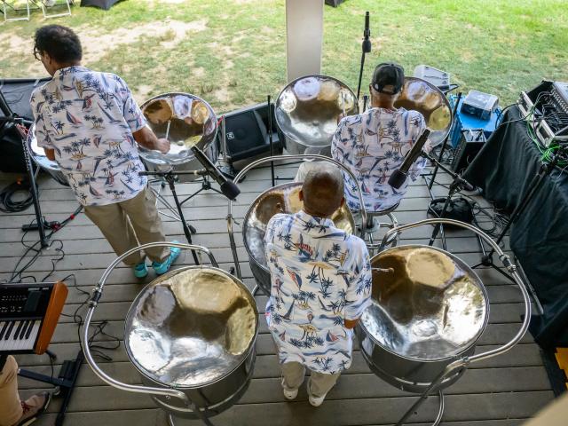 Three musicians wearing patterned shirts play steel drums on a wooden outdoor stage, surrounded by audio equipment, with grass visible in the background.