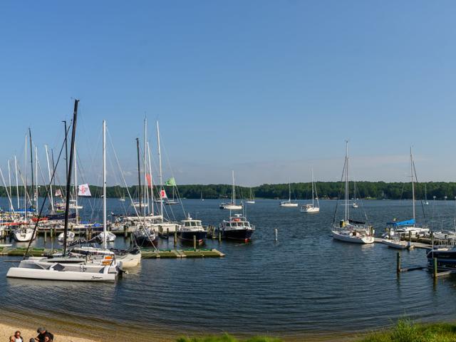 Panoramic view of a marina with multiple sailboats docked, a small sandy beach with kayaks in the foreground, and calm water surrounded by greenery under a clear blue sky.