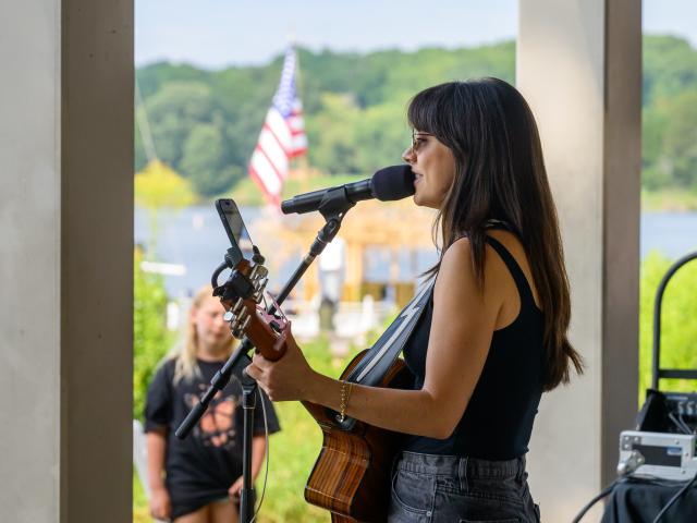 Woman playing acoustic guitar and singing into a microphone at an outdoor venue, with an American flag and greenery visible in the background.
