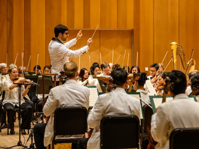 A conductor leads an orchestra of musicians dressed in white jackets, performing on stage with various instruments and a wooden background.