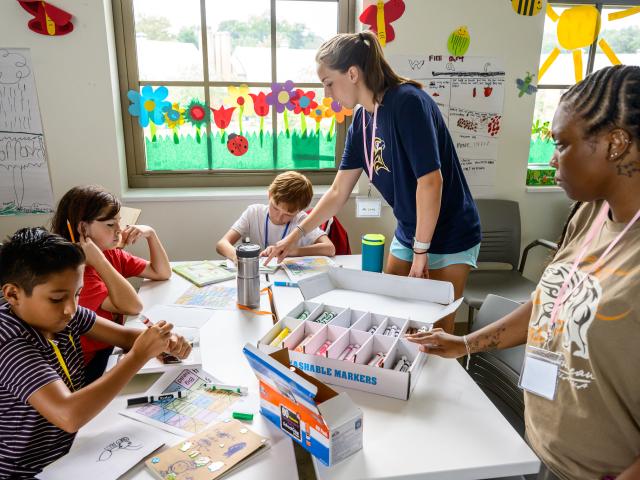 Three children sit at a table working on crafts with markers as two adults supervise in a bright classroom decorated with colorful art and paper bees.