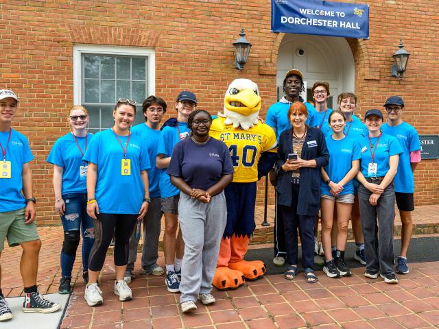 A group of people in blue shirts pose with a mascot in front of Dorchester Hall, standing on a brick path by a building entrance.