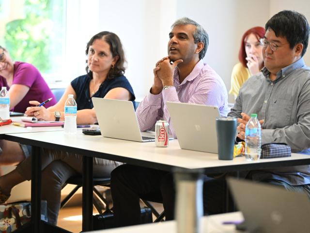 Five adults sit at tables with laptops, notebooks, and drinks, appearing attentive and engaged in a meeting or classroom setting.