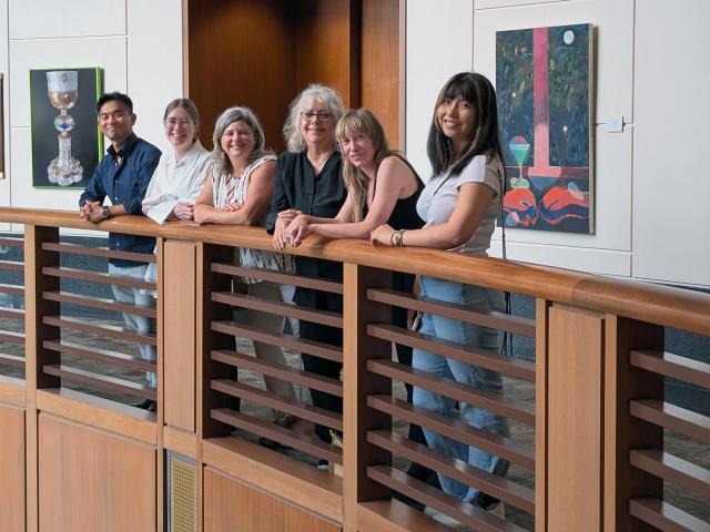Six people stand in a row, leaning against a wooden railing inside a building with art displayed on the walls behind them.