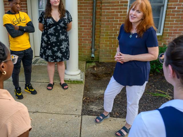 A woman speaks to a small group of people standing in a semicircle outside a brick building with a white pillar and window.