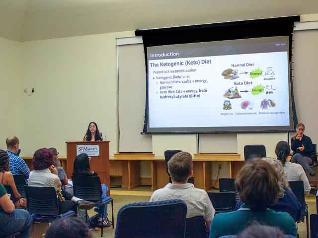 A woman presents a slideshow about the ketogenic diet to an audience in a classroom; a sign language interpreter sits to her right.