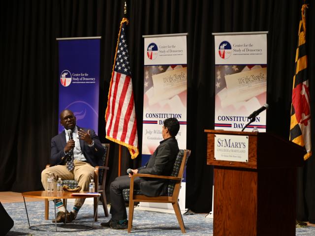 Two people sit on stage having a discussion during a Constitution Day event at St. Mary's College of Maryland, with banners and an American flag in the background.