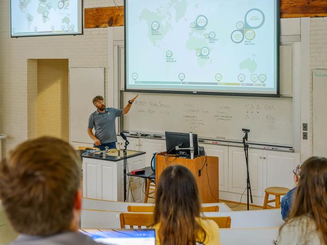 A man stands at the front of a classroom, pointing at a world map on a projector screen, while students sit facing him and take notes.