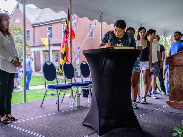 A student signs a book on a podium while others wait in line; an adult stands to the side on a stage set up outdoors with chairs and flags visible.