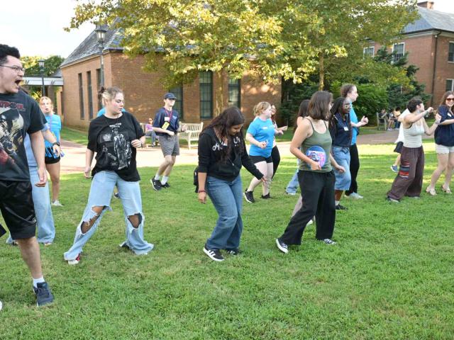 A group of people stand on a lawn, participating in a coordinated outdoor activity or dance, with buildings and trees in the background.
